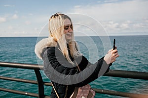 A beautiful smiling blonde girl makes a selfie on the phone against the background of the sea.