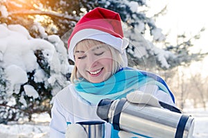 Beautiful smiling blonde girl in a red Christmas hat pours hot tea into a cup