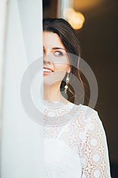 Beautiful smiling bride brunette young woman in white lace dress near window, close up