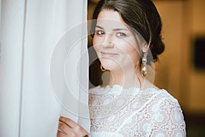 Beautiful smiling bride brunette young woman in white lace dress near window, close up portrai