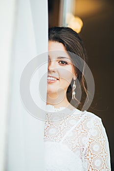 Beautiful smiling bride brunette young woman in white lace dress near window, close up portrait