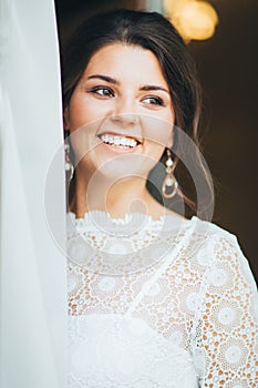 Beautiful smiling bride brunette young woman in white lace dress near window, close up portrait