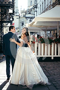 Beautiful smiling bride is hugging with the groom while walking along the sunny street with the wedding bouquet. Back