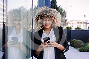 Beautiful smiling business woman using mobile phone and headphones in city. Buildings background