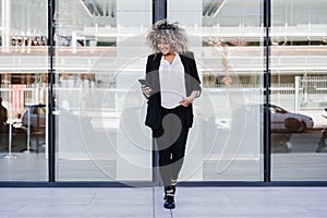 Beautiful smiling business woman using mobile phone and headphones in city. Buildings background