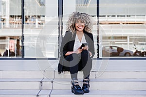 Beautiful smiling business woman using mobile phone sitting on stairs in city. Buildings background