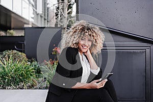 Beautiful smiling business woman using mobile sitting on stairs in city. Buildings background