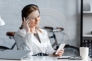 Beautiful smiling businesswoman in headphones sitting at desk and using smartphone