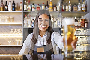 Beautiful smiling female Bartender serving a draft beer at the bar counter , shelves full of bottles with alcohol on the