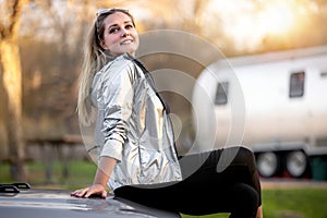 A beautiful smiling female traveler sitting on car in campground with RV trailer in background, sunset golden hour