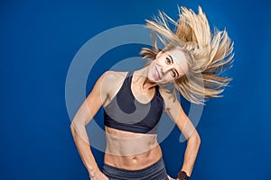 Beautiful smiling fit young woman in sport bra with flowing hair in studio on a blue background. Dance, gym, slim concept