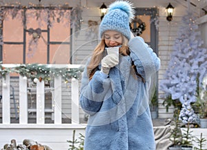 Beautiful smiling girl drinking tea, coffee or other hot drink in winter on the background of a christmas house