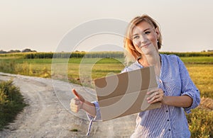 Beautiful smiling girl gets hitchhiked and votes with a sign of cars on a rural road in the rays of the sunset