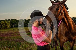 Beautiful smiling girl jockey stand next to her brown horse wearing special uniform on a sky and green field background