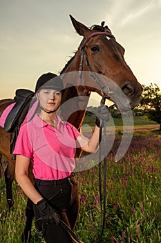 Beautiful smiling girl jockey stand next to her brown horse wearing special uniform on a sky and green field background