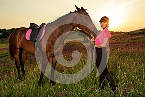Beautiful smiling girl jockey stand next to her brown horse wearing special uniform on a sky and green field background