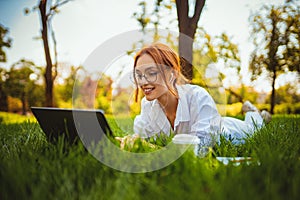 beautiful smiling girl student uses laptop pc while lying on the grass outdoors. Education concept