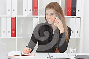 Beautiful smiling girl talking on the mobile phone sitting at office and looking at laptop.