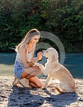 Beautiful smiling girl training her golden retriever puppy on sandy beach near the river enjoying their connection.