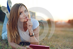 Beautiful smiling girl in white T-shirt and jeans lying in the grass on the field and holding a tab in her hand