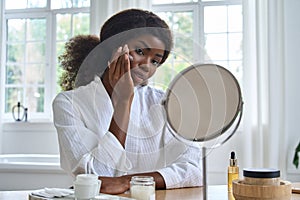 Young happy black girl applying facial cream in front of mirror in bathroom.