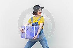 Beautiful smiling hipster woman with bright makeup in blue denim overalls holding big present box.