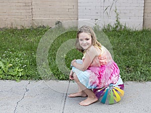 Beautiful smiling little girl in summer dress and bare feet sitting on beach ball on sidewalk