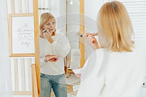 Beautiful smiling mature senior caucasian woman with blonde hair standing near mirror with make up palette and brush