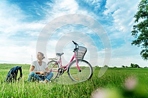 Beautiful smiling mature woman sitting next to bike on the grass on a green field. Summer Country Vacation Adventure Concept