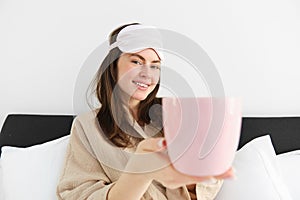 Beautiful smiling modern woman in pyjamas and sleeping mask, offering cup of tea or coffee in bed and looking happy