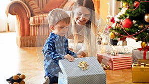 Beautiful smiling mother looking at her boy opening Christmas gifts and presents