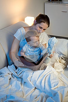 Beautiful smiling mother reading fairytale book to her baby at b