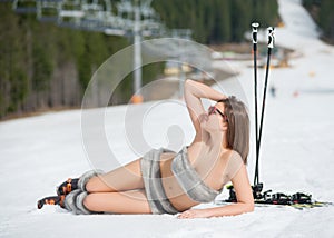 Beautiful smiling naked skier is lying on snowy slope near ski lift at ski resort