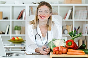 Beautiful smiling nutritionist looking at camera and showing healthy vegetables in the consultation.