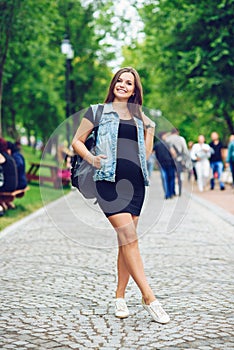 Beautiful smiling pregnant woman in a black tight dress and blue jeans jacket with backpack walking on the alley in the