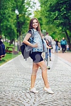 Beautiful smiling pregnant woman in a black tight dress and blue jeans jacket with backpack walking on the alley in the