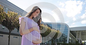 Beautiful smiling pregnant woman standing outdoor and stroking her belly. Concept of happy pregnancy and baby