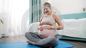 Beautiful smiling pregnatn woman in sportrs outfit sitting on fitness mat and stroking her big belly. Concept of healthy