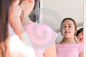 Beautiful smiling preteen girl looking at her reflection in mirror while her mother doing hair