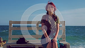 Beautiful smiling red-haired traveler girl sitting on a bench on the sea beach, dreams, relaxes and enjoys life.