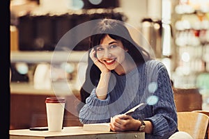 Beautiful smiling student woman reading a book in the cafe with warm cozy interior and drinking coffee