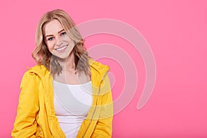 Beautiful smiling trendy teenage girl in bright yellow jacket looking at camera. Attractive young woman portrait over pastel pink.