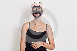 Beautiful smiling woman with black clay facial mask on face standing against white wall with charming smile, cute girl doing