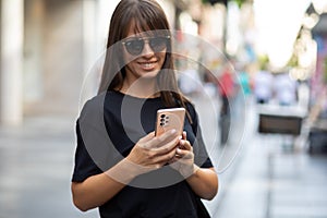 Beautiful smiling woman in black shirt and sunglasses texting on smartphone on street in city center