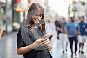 Beautiful smiling woman in black shirt texting on smartphone on street. Blurred people in background