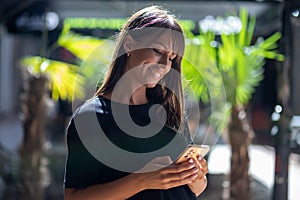 Beautiful smiling woman in black shirt texting on smartphone on street in city center. Palm in background