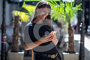 Beautiful smiling woman in black shirt texting on smartphone on street in city center. Palm in background