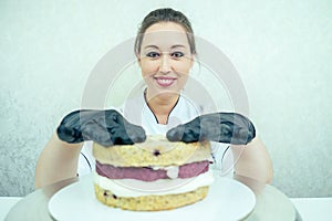 A beautiful and smiling woman confectioner in black gloves and a white working uniform bakes a cake in the kitchen