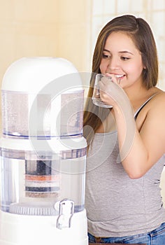 Beautiful smiling woman drinking a glass of water with a filter system of water purifier on a kitchen background