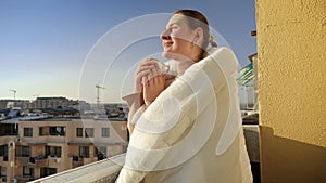 Beautiful smiling woman drinking tea on the terrace and looking at sunrise over the city. People in morning, relaxing at home.